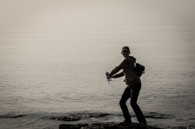 A person standing on a rocky shoreline holding a water bottle, silhouetted against a calm and misty body of water, evoking a serene and adventurous mood.