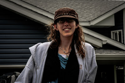 A woman with wavy hair wearing a brown hat and glasses, smiling outdoors in front of a house with blue siding and white trim.